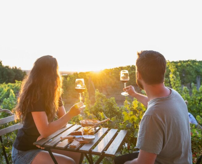 couple à table avec des verres de vin à la main face au coucher du soleil devant des vignes